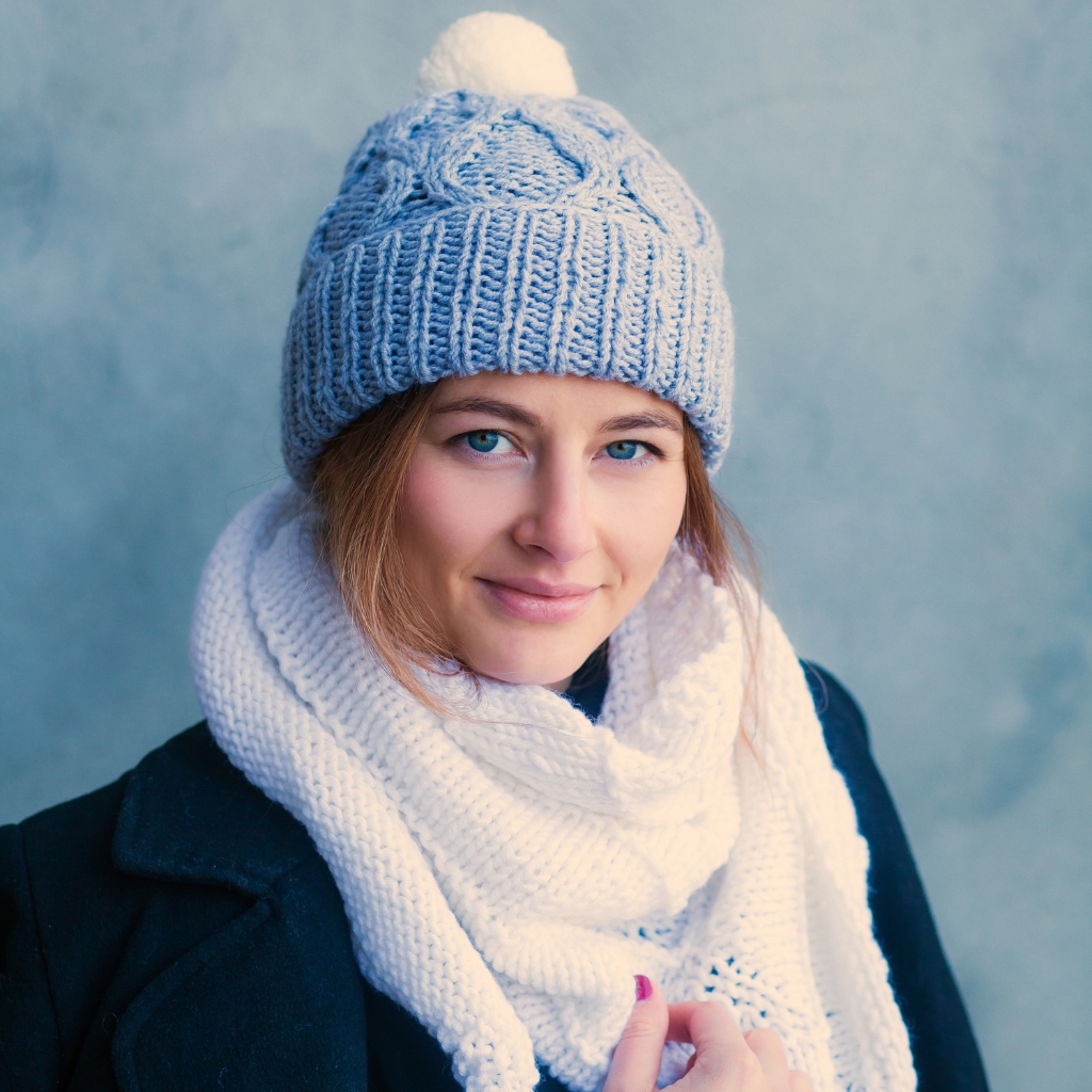 A woman wearing a chunky blue knit beanie with a white pom-pom and a white scarf, standing against a pale blue background.