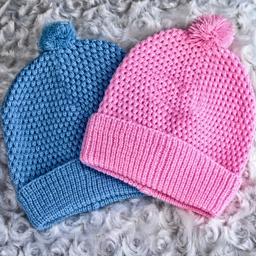 A pair of handmade crochet beanies: one blue and one pink, each with a textured waffle stitch and a fluffy pom-pom on top, laid on a soft, light-gray faux fur surface.