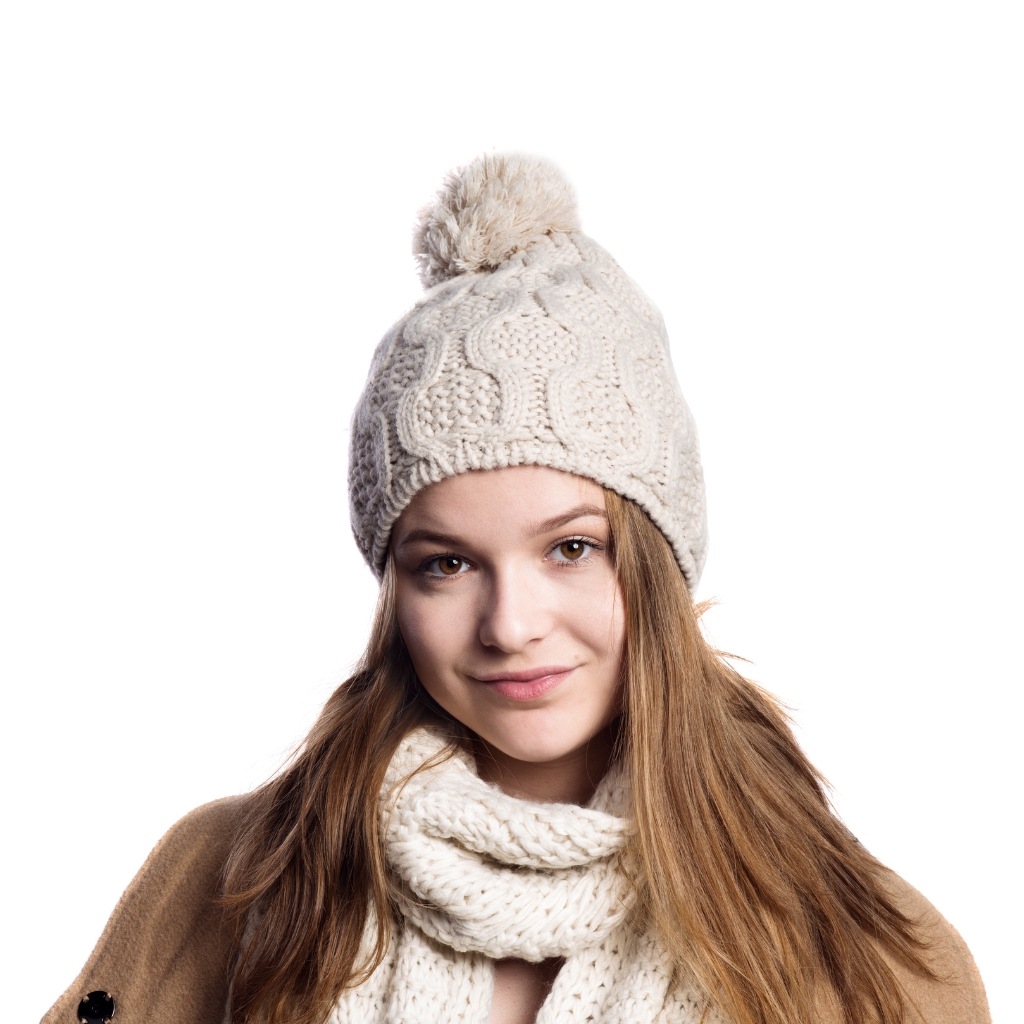 A young woman wearing a chunky white cable-knit beanie with a fluffy pom-pom, scarf, and camel coat, posing against a white background.