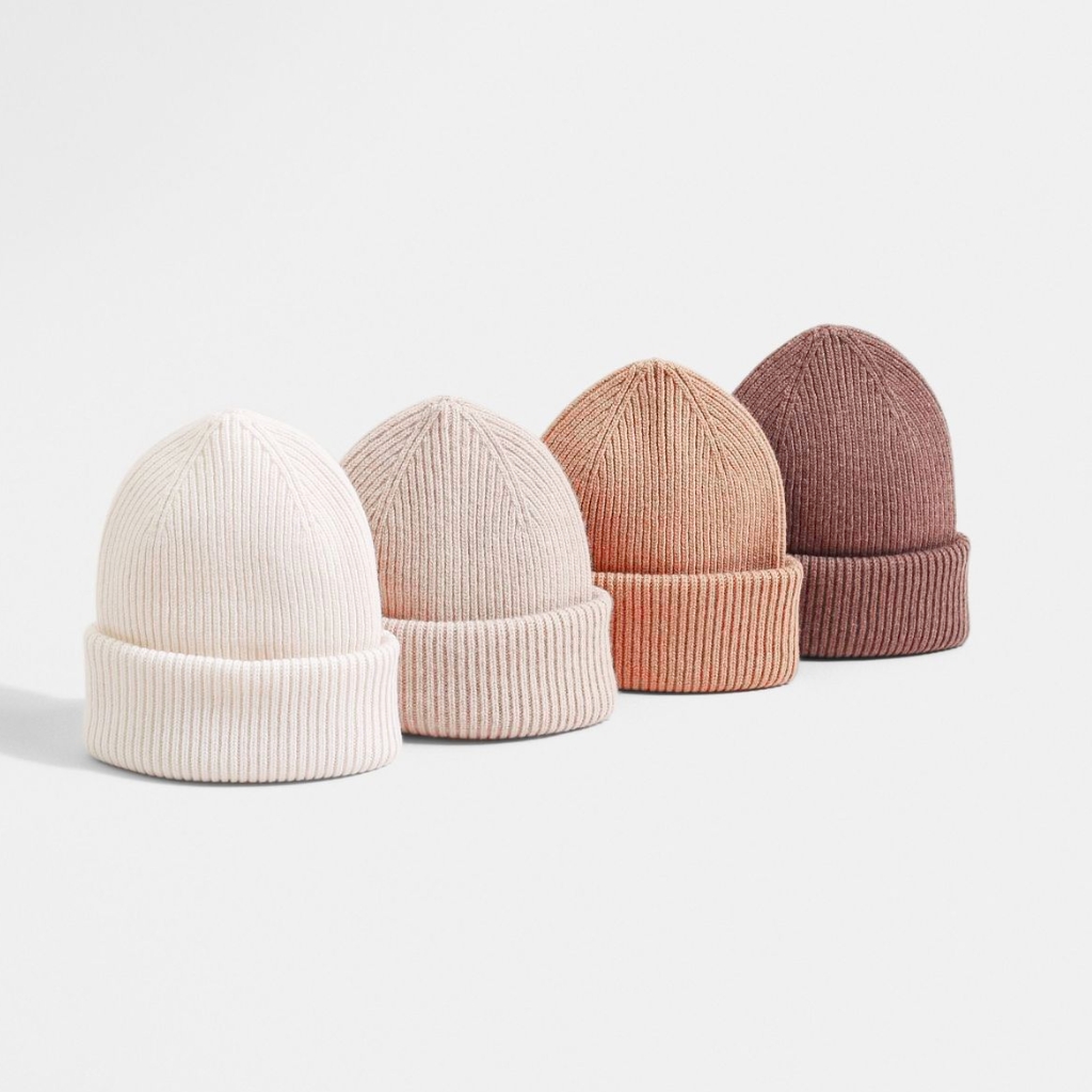 Four knit beanies in a row—creams and neutrals fading from left to right (off-white, light beige, soft peach, dusty mauve)—each with a folded brim and ribbed texture, displayed against a pale background.