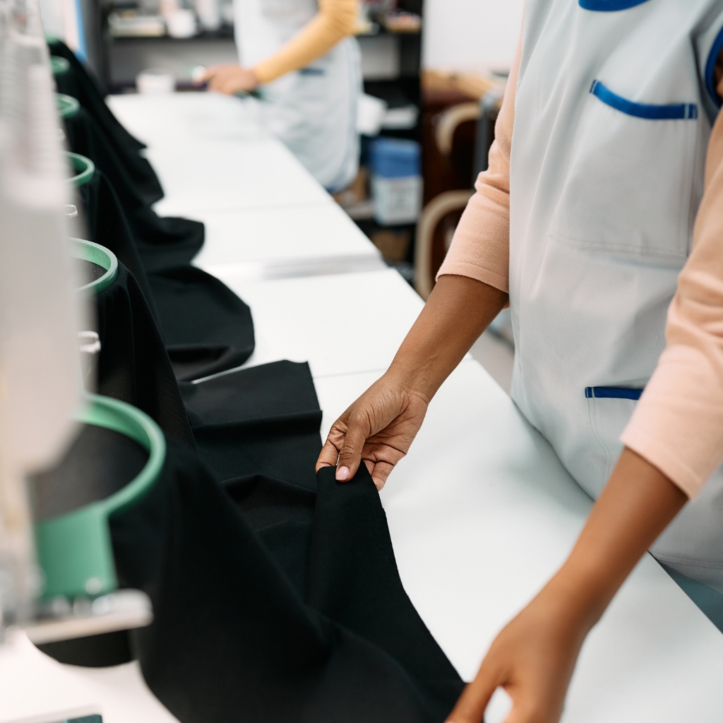 A close-up of a seamstress or worker at a sewing station, wearing a light-colored uniform with blue trim, carefully handling black fabric on a white table. Industrial sewing equipment and additional fabric pieces are visible in the background.