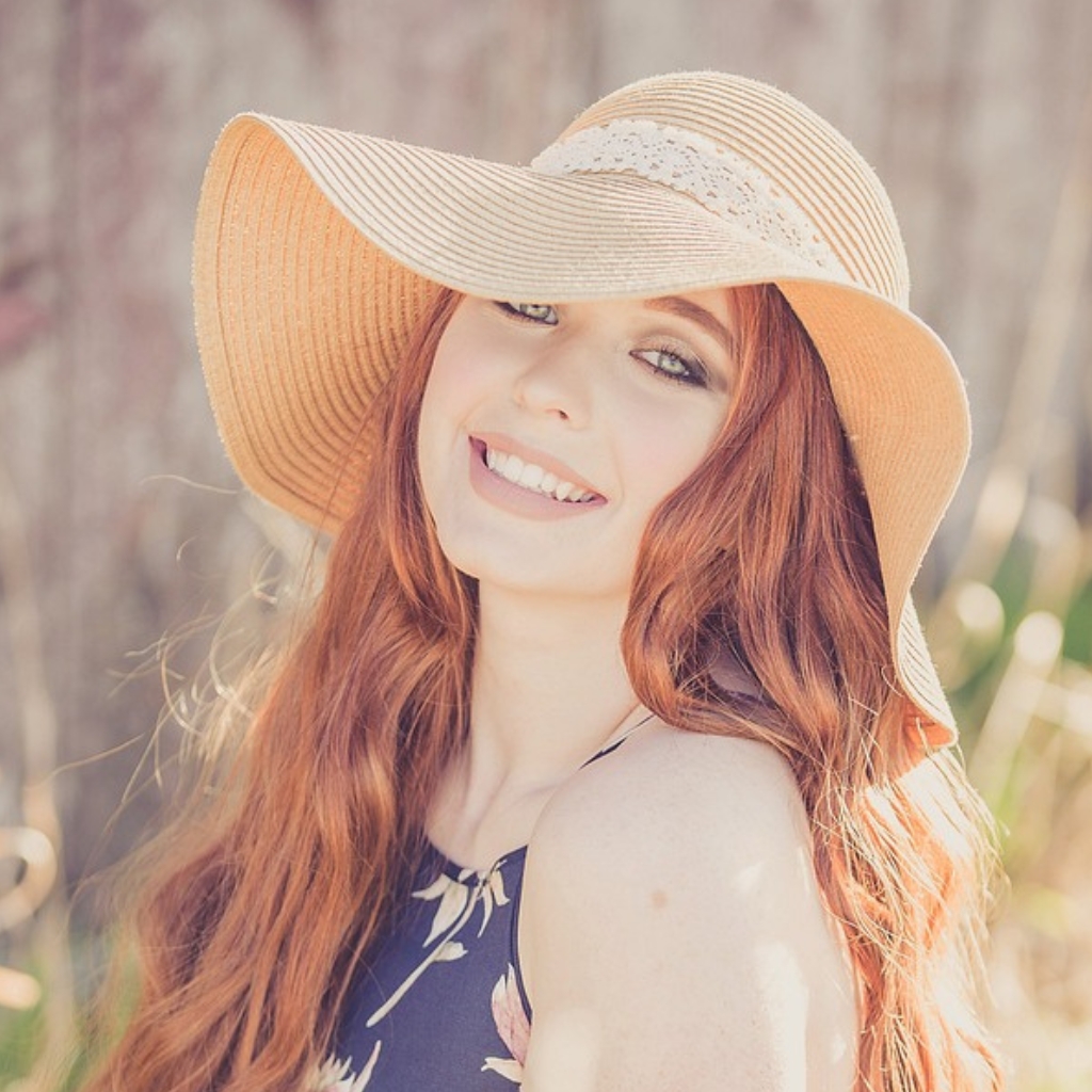 A smiling woman with long red hair wearing a large, floppy straw sun hat outdoors, bathed in warm sunlight.

