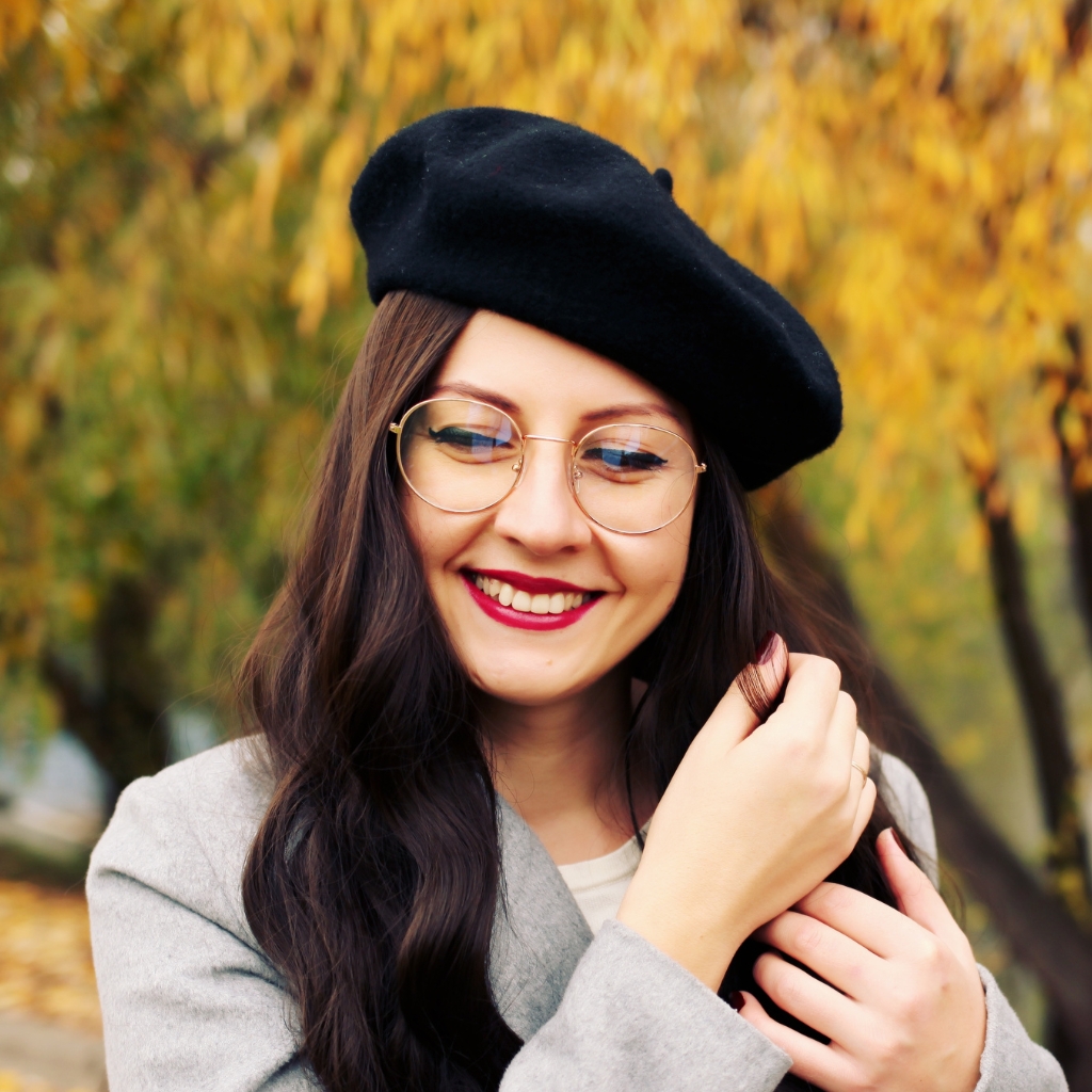 Une femme souriante portant un béret noir, des lunettes rondes, du rouge à lèvres rouge et un manteau léger, à l'extérieur avec un feuillage automnal en arrière-plan.