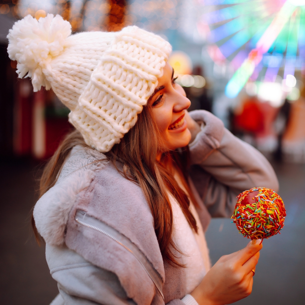 A smiling woman wearing a chunky white knit beanie with a pom-pom, holding a colorful candy-coated cake pop with rainbow sprinkles. She is dressed in a light coat in an outdoor festive setting with blurred lights in the background.