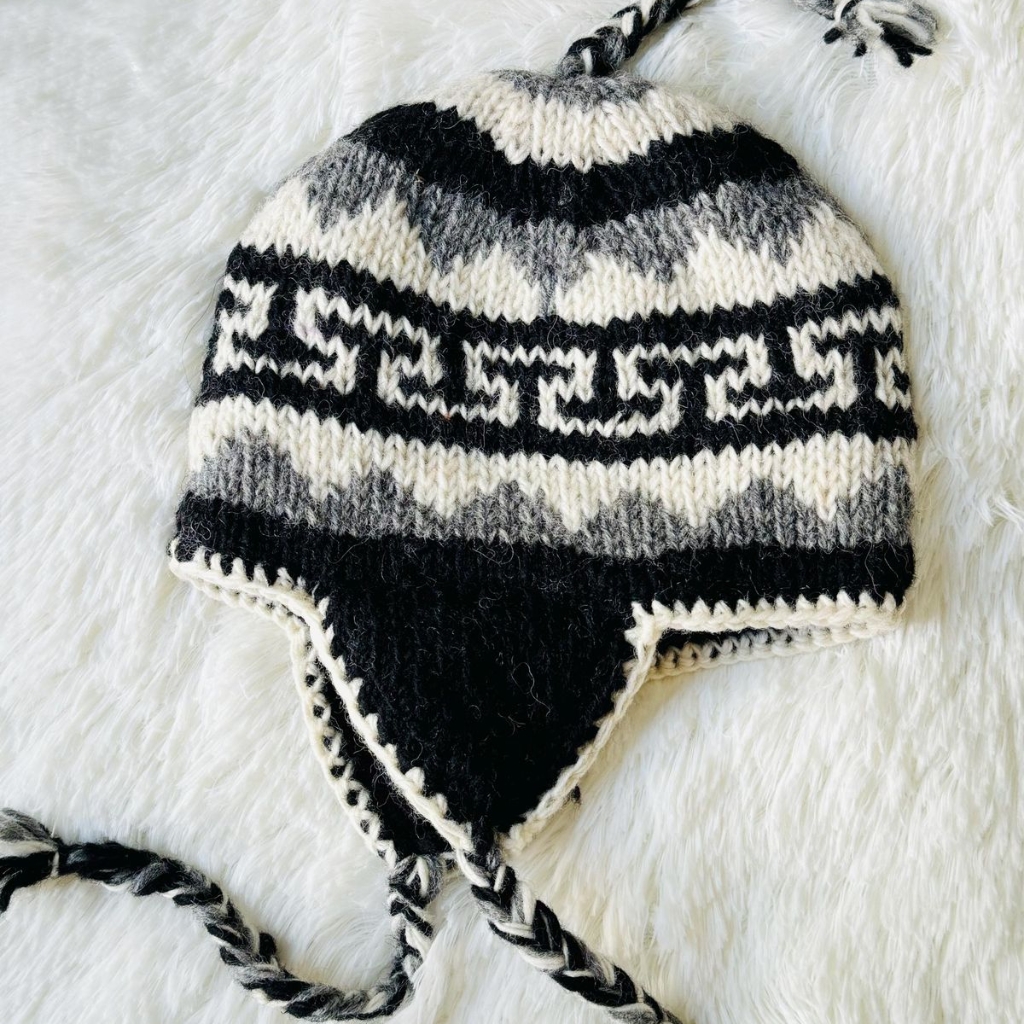 Un gorro de invierno de punto negro, blanco y gris con orejeras y cordones trenzados, con un diseño geométrico, sobre una superficie blanca suave.