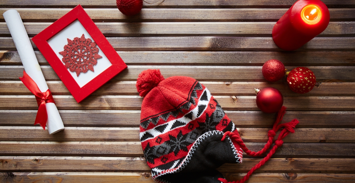 Eine festliche Winterszene auf einer Holzoberfläche mit einer rot-grauen Strickmütze mit Ohrenklappen, umgeben von roten Weihnachtskugeln, einer roten Kerze, einem gerollten Weihnachtsgutschein, der mit einem roten Band zusammengebunden ist, und einer rot gerahmten Schneeflockenverzierung.