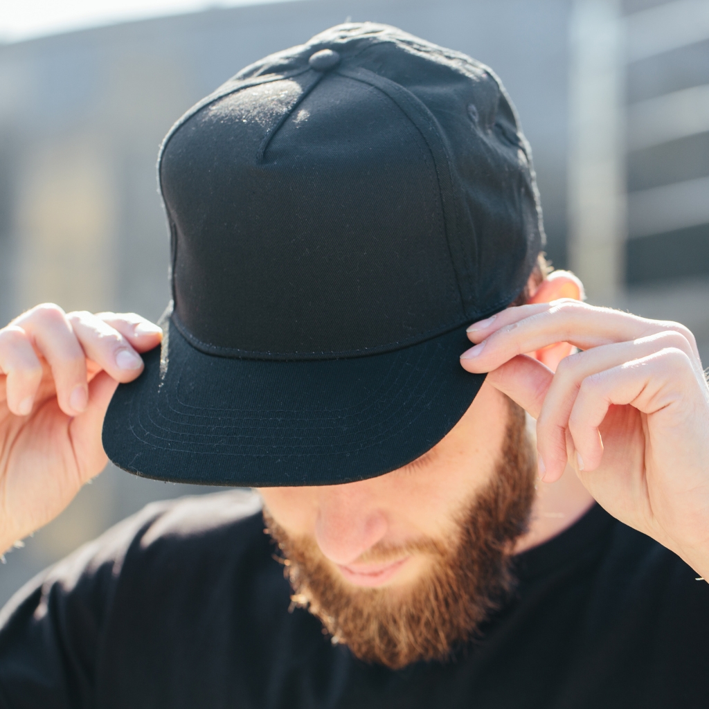 Nahaufnahme eines Mannes, der eine schlichte schwarze Baseballkappe trägt und deren Schirm mit beiden Händen nach unten drückt, während er sie zurechtrückt. Er hat einen Bart und trägt ein schwarzes Hemd, im Hintergrund ist eine urbane, sonnenbeschienene Kulisse zu sehen. 