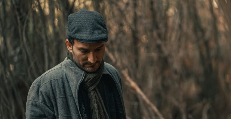 Ein Mann mit Schirmmütze und Schal steht vor einem gedämpften, bewaldeten Hintergrund und blickt nach unten.