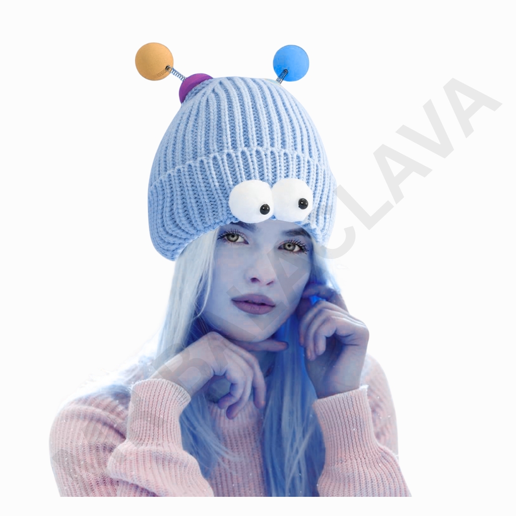 A person wearing a chunky blue knit hat with two large white googly eyes on the front and colorful antennae on top, posing with hands near the face.