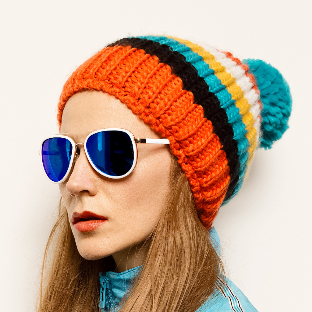Eine Frau mit einer bunten Strickmütze und einer blauen Sonnenbrille mit verspiegelten Gläsern, die leicht nach links blickt.