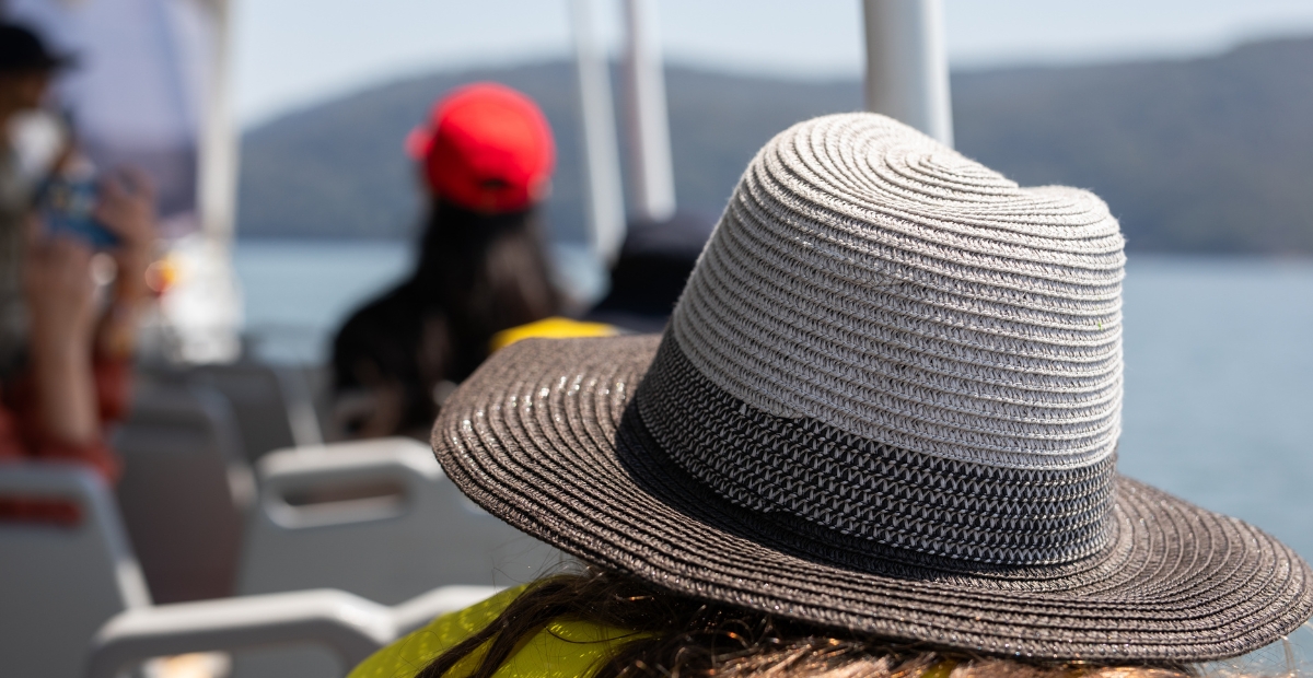 Nahaufnahme eines breitkrempigen Strohhutes mit einem schwarzen Band, getragen von einer Person auf einem Boot, verschwommene Passagiere und Wasser im Hintergrund im