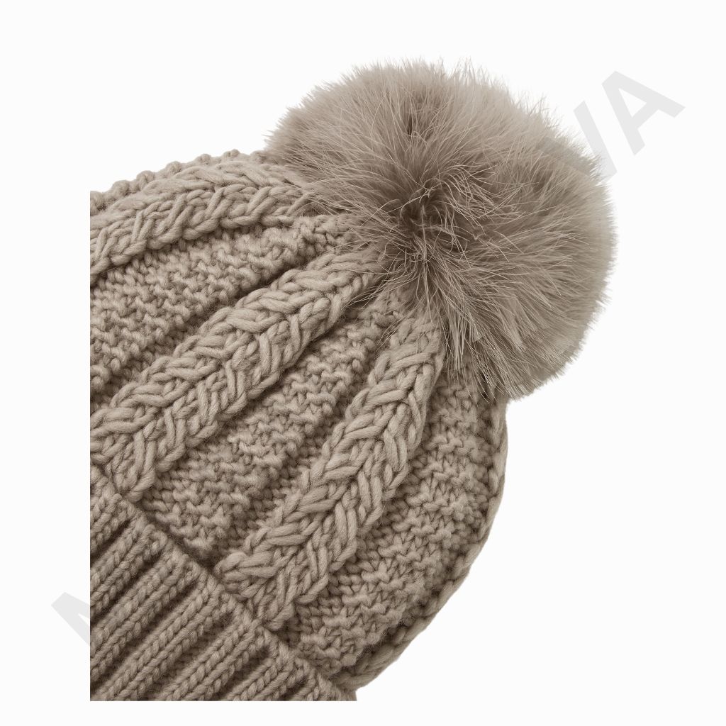 A close-up of a beige knitted beanie with a large, soft pom-pom on top, featuring chunky cable and ribbed knit patterns.