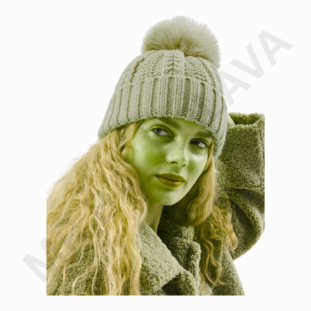 A portrait of a person with long wavy hair wearing a chunky knit beanie with a fluffy pom-pom and a textured coat. The image has a green tint.