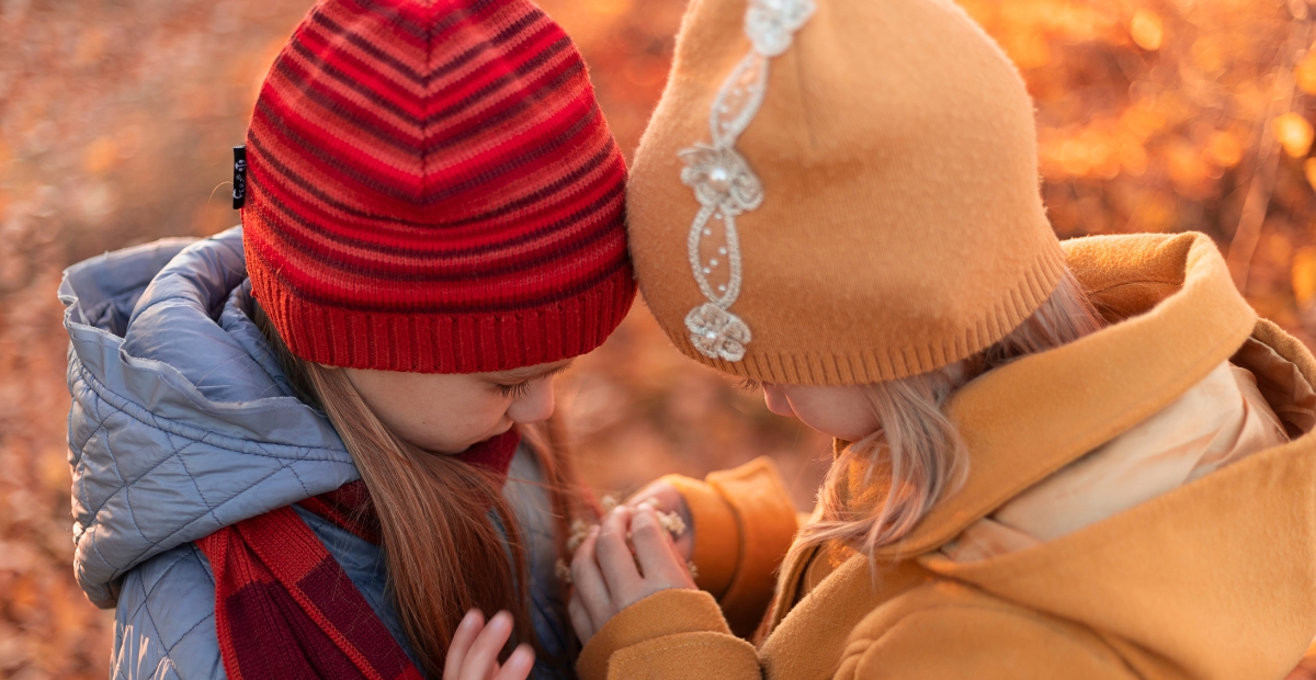 Zwei Kinder mit doppellagigen Strickmützen, eine rot gestreift und eine orange mit einer dekorativen Anstecknadel oder einem Anhänger, lehnen sich in einer herbstlichen Umgebung eng aneinander.