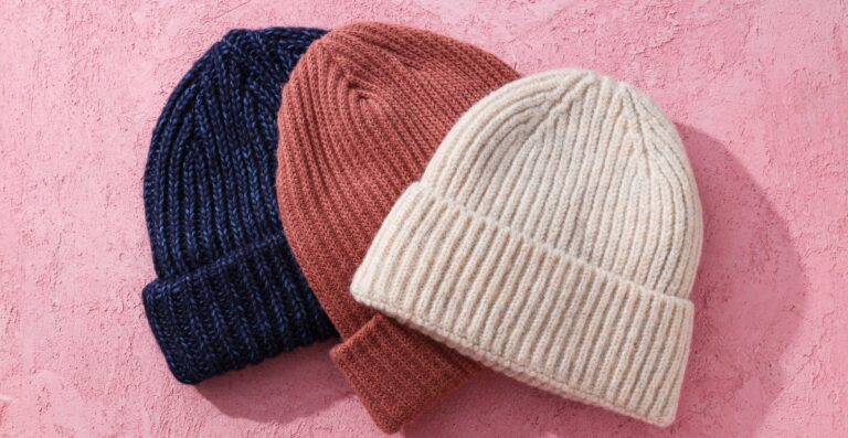 Drei Strickmützen in Marineblau, Altrosa und Creme auf einem rosa strukturierten Hintergrund angeordnet.