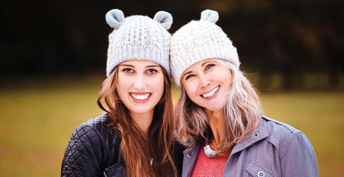 Zwei Frauen mit originellen Strickmützen mit kleinen Ohren, die vor einem verschwommenen grün-braunen Hintergrund im Freien lächeln.
