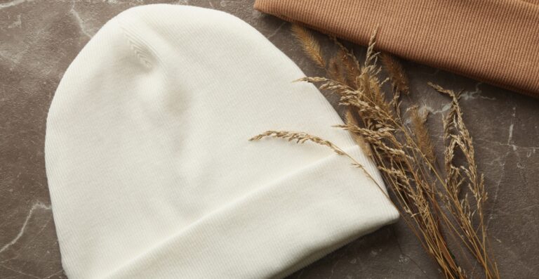 Nahaufnahme einer weißen Strickmütze und einer hellbraunen Strickrolle, neben denen getrocknete Gräser auf einer strukturierten Oberfläche liegen.