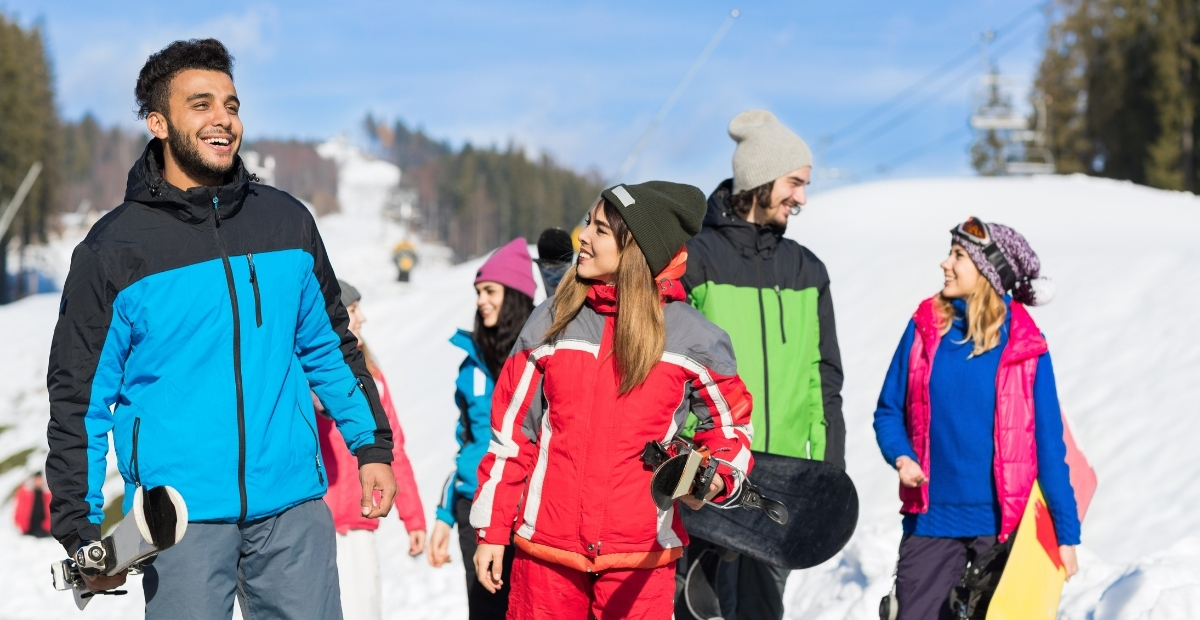Eine Gruppe von Freunden in bunten Skijacken läuft auf einem verschneiten Hang, lächelt und trägt Snowboards und Skiausrüstung.