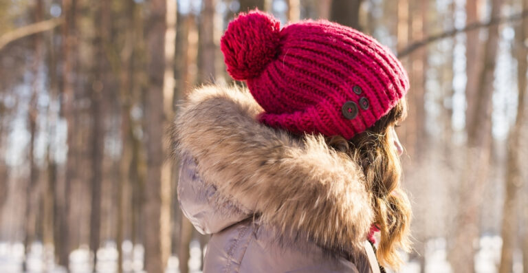 Eine Person, die eine leuchtend rosa Strickmütze mit einem flauschigen Bommel trägt und mit einer pelzgefütterten Kapuze in einem verschneiten Wald steht.