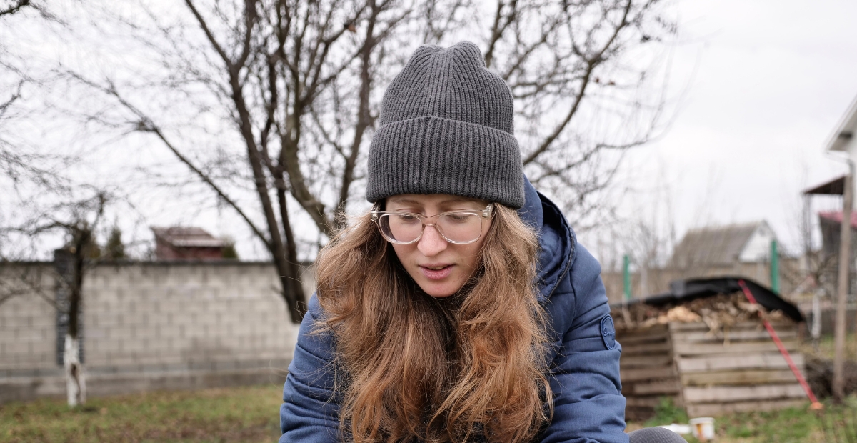 Eine junge Frau im Freien, die eine graue Strickmütze, eine runde Brille mit klarem Rahmen und eine blaue Daunenjacke trägt. Sie hat langes, welliges Haar und schaut nach unten, im Hintergrund sind ein kahler Baum und ein Wohngebiet zu sehen.