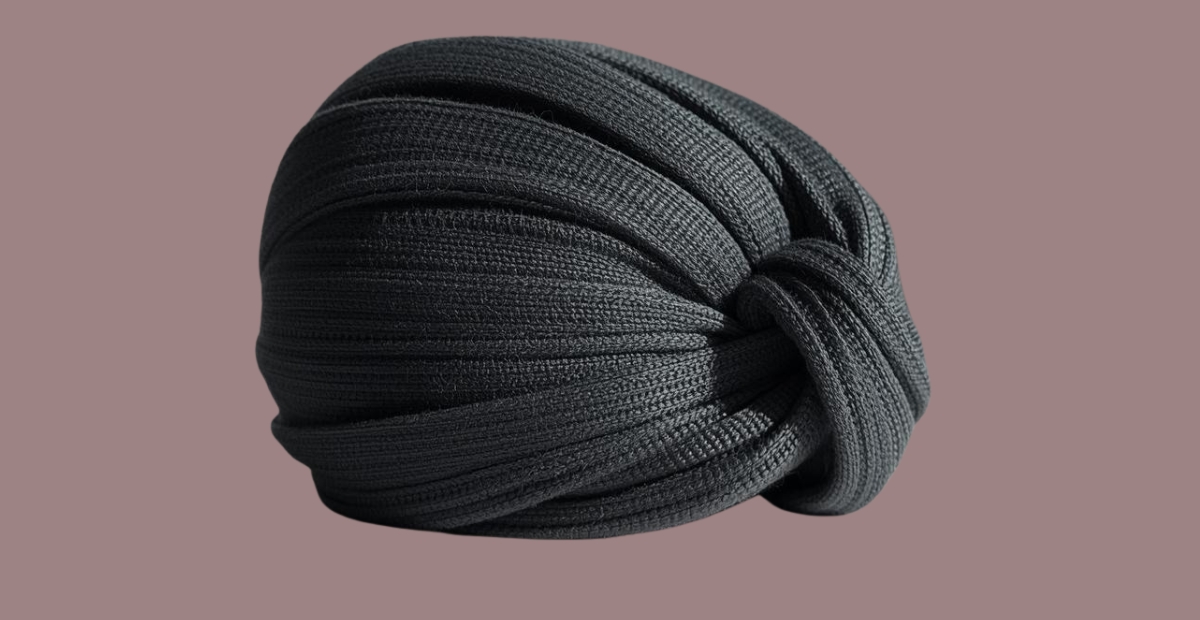Eine dunkelgraue Strickmütze im Turban-Stil mit mehreren gedrehten Bändern, die an der Seite einen Knoten bilden, vor einem gedeckten mauvefarbenen Hintergrund.