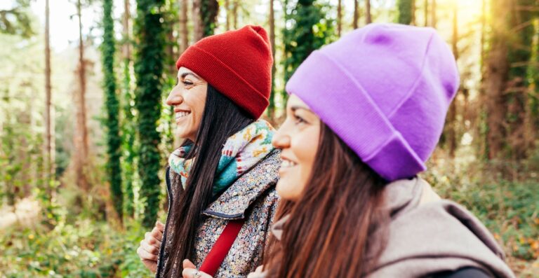 Zwei Frauen, die in einem Wald spazieren gehen, tragen bunte vegane Mützen (rot und lila), lächeln und genießen die Natur.