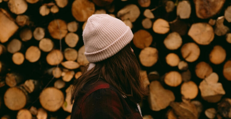 Eine Person mit langen dunklen Haaren, die eine beige Strickmütze und eine dunkle karierte Jacke trägt und vor einem großen Stapel geschnittener Holzscheite steht.