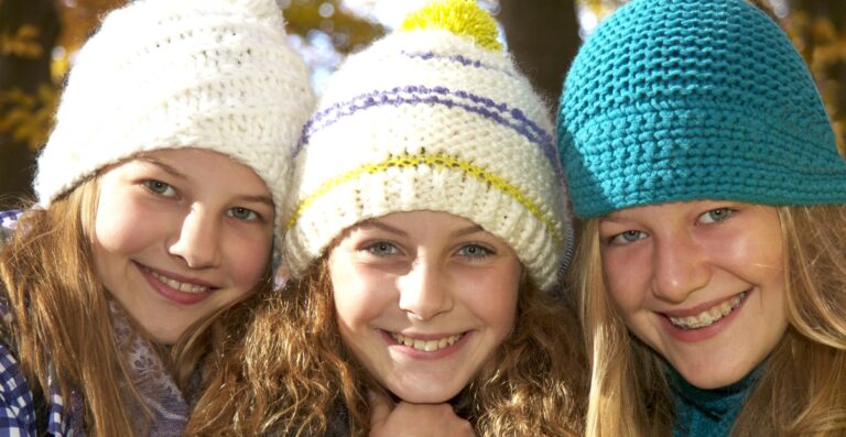 Drei lächelnde Mädchen mit dicken Waffelstrickmützen: links eine weiße, in der Mitte eine weiße mit bunten Streifen und rechts eine türkisfarbene, draußen im Herbstlicht.