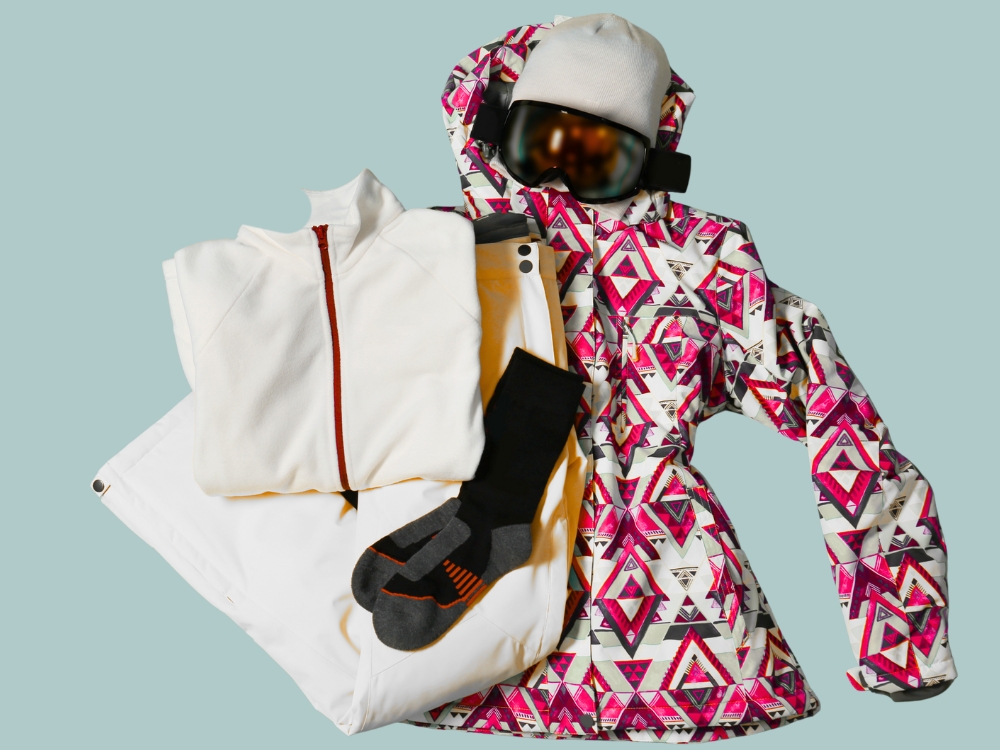A ski wear set laid out with a pink geometric-patterned jacket and matching pants, a white insulated outer layer, black gloves, and orange accents, designed for cold-weather snowsports