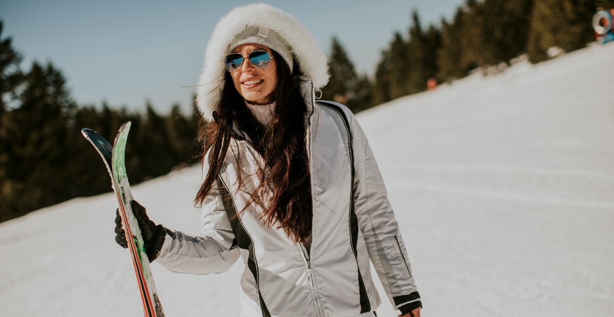 Eine Frau auf einem verschneiten Hang, die eine weiße Winter-Skijacke mit pelzgefütterter Kapuze und eine reflektierende Sonnenbrille trägt, Skier in den Händen hält und lächelt. Im Hintergrund sind immergrüne Bäume und ein klarer blauer Himmel zu sehen.