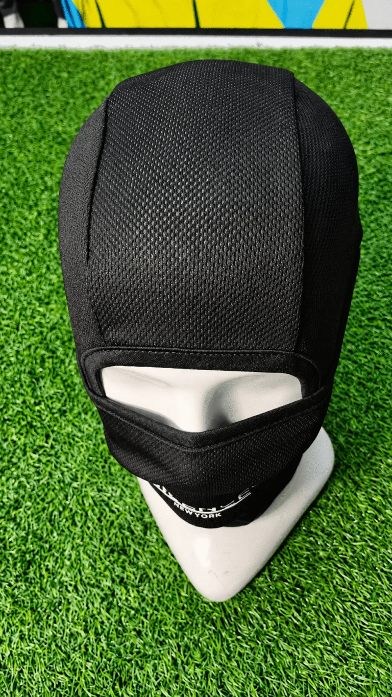 Black balaclava with mesh texture across the upper head, covering a white mannequin head and neck with separate eye, nose, and mouth openings. White text reading "FCE NEW YORK" runs along the lower section, against a blurred green turf background with yellow and light blue shapes.