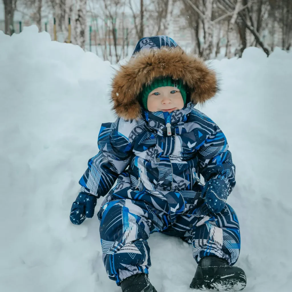 Small child sitting in the snow in a printed one-piece snowsuit with a fur-trimmed hood